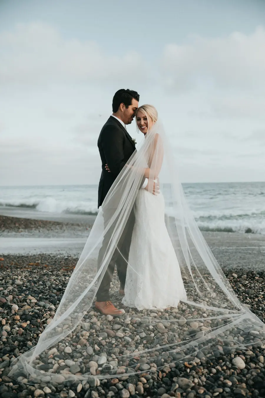 Bride and groom embracing on the beach, her veil drifting in the breeze
